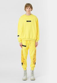Sudadera amarilla y joggers, con un parche de logo negro y un diseño en forma de pluma en los pantalones. La tela parece suave y con un ajuste relajado.