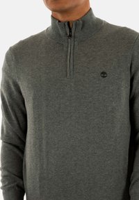 Maglione grigio a maglia con colletto con zip, polsini a coste e un piccolo logo sul petto. Il tessuto sembra morbido e testurizzato.