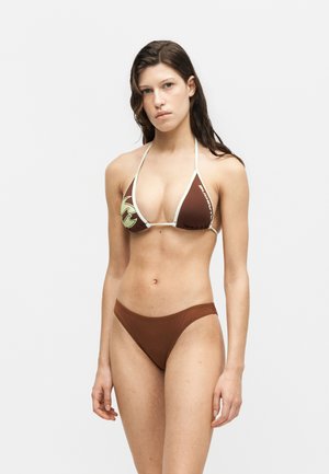 Fiatal nő barna és krémszínű háromszög alakú bikini felsővel és hozzáillő barna bikini alsóval, sima világos háttér előtt áll.