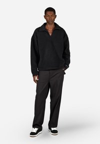 Čierny fleecový pulóver so širokým golierom, textúrovaným povrchom a voľným strihom, skombinovaný s tmavými nohavicami a čierno-bielymi teniskami.