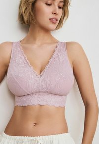 Bralette in pizzo rosa con design a scollo a V, bordi ondulati e una vestibilità aderente sotto il busto. Il tessuto presenta un motivo floreale e una consistenza morbida.