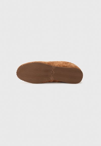 Semelle de chaussure en daim marron avec un motif de tread en zigzag, surface texturée et forme arrondie. Design minimaliste sans aucune marque visible.