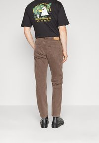 Mies, jolla on päällään ruskeat farkut, mustat saappaat ja musta T-paita, jossa on värikäs grafiikka ja teksti takana.