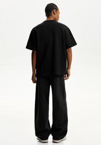 Tricou negru supradimensionat cu mâneci scurte, realizat dintr-un material moale. Persoana poartă blugi negri largi și teneși de culoare deschisă.