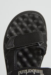 Svart sandal med en texturerad, mönstrad fotbädd, justerbara remmar och en slitstark sula. Kardborrefäste för säker passform.