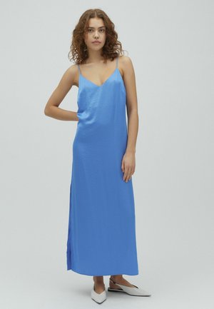 Młoda kobieta o kręconych włosach, ubrana w niebieską sukienkę maxi bez rękawów i białe spiczaste baleriny na pasku, stoi z jedną ręką na biodrze.