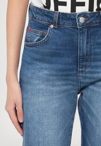 Gros plan d'une personne portant un jean bleu taille mi-haute avec des coutures visibles et un bouton à l'avant, main reposant sur le côté.