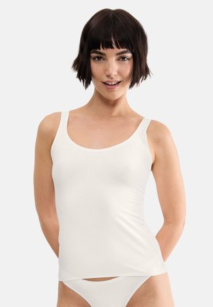 Kobieta o krótkich ciemnych włosach, ubrana w biały prążkowany top na ramiączkach i dopasowaną białą bieliznę na jednolitym jasnym tle.