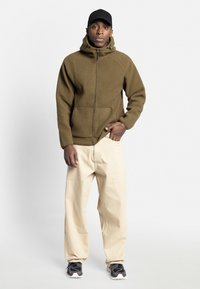Fleecejakke i olivengrøn med lynlås foran, hætte og sidelommer. Sammen med beige bukser og sorte sneakers med detaljer.