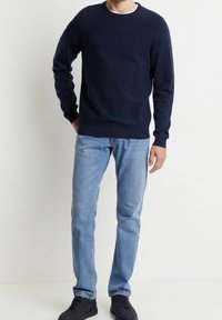 Mörkblå tröja med rund halsringning, tillverkad av mjukt material, tillsammans med ljusblå jeans och svarta sneakers, stående mot en vit bakgrund.