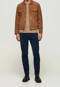 Man som bär en brun mockajacka, en beige kabelstickad tröja, mörkblå byxor och svarta läderstövlar som står framför en enfärgad bakgrund.