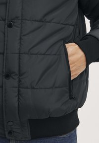 Giacca imbottita nera con chiusura a bottoni frontale, tasca laterale con zip e polsini a coste per un maggiore comfort. Tessuto liscio con design trapuntato.