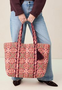 Handen die een grote doorgestikte tas vasthouden met een roze, groene en kastanjebruine bloemmotief, gedragen met een blauwe spijkerbroek, een bordeauxrode trui en schoenen met studs.