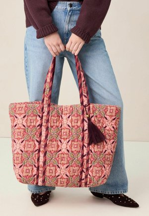 Hände halten eine große gesteppte Tragetasche mit rosa, grünen und kastanienbraunen Blumenmuster, getragen zu blauer Jeans, burgunderfarbenem Pullover und mit Nieten verzierte Schuhe.