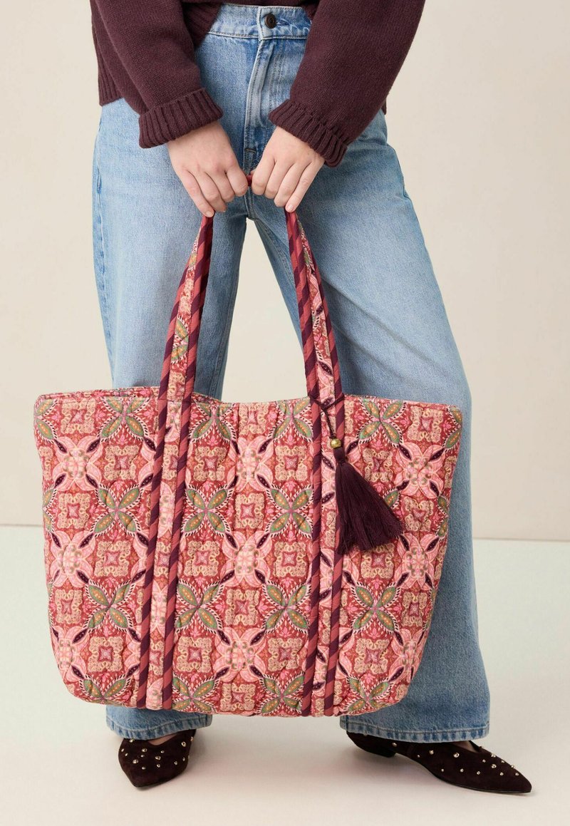 Handen die een grote doorgestikte tas vasthouden met een roze, groene en kastanjebruine bloemmotief, gedragen met een blauwe spijkerbroek, een bordeauxrode trui en schoenen met studs.