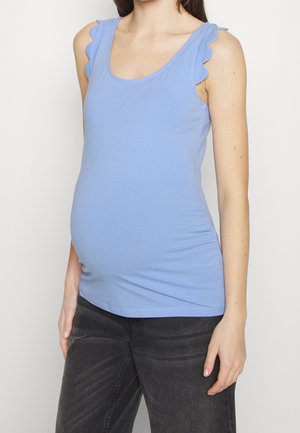 Kobieta w ciąży w jasnoniebieskiej bluzce na ramiączkach z falistymi detalami na ramionach oraz ciemnoszarych spodniach, stojąca na białym tle.