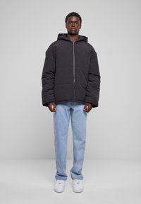 Sort polstret jakke med hætte, med en frontlynlås og brede ærmer, bæres over lys blå jeans og hvide sneakers.