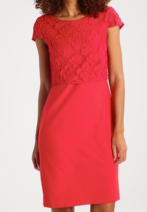Kobieta w dopasowanej czerwonej sukience z kwiatowym teksturowanym krótkim rękawem na górze i gładką spódnicą na białym tle.
