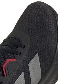 Gros plan d'une chaussure de sport noire avec tissu texturé, lacets noirs, détail rouge près de la zone des lacets, et deux bandes grises sur le côté.