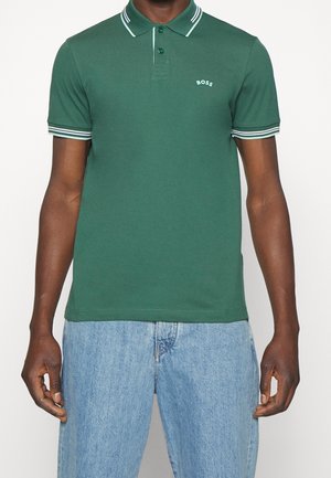 Man wearing a green polo shirt with white stripes on collar and sleeves, paired with light blue denim jeans.