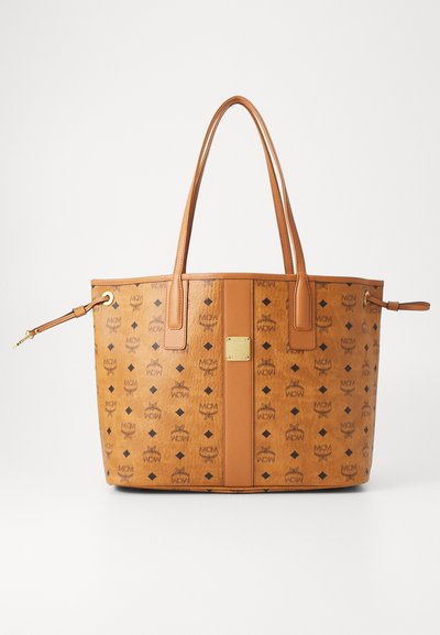 Tan leather tote bag with black MCM logo pattern, two handles, side straps, and a centered gold metal brand plaque.