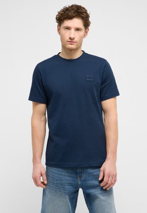 Mann mit lockigem Haar, der ein dunkelblaues kurzärmeliges T-Shirt und hellblaue Jeans trägt, steht vor einem schlichten hellen Hintergrund.