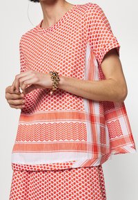 Žena v oranžovém a bílém vzorovaném topu s krátkým rukávem a odpovídající sukni, se zlatým perličkovým náramkem na levém zápěstí.