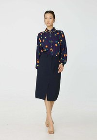 Blouse bleu marine avec un imprimé floral coloré, devant à boutons et manches bouffantes, associée à une jupe sombre longueur genou avec une fente frontale.
