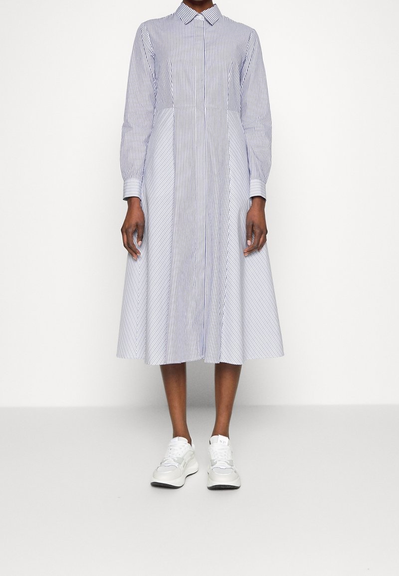 Robe chemise rayée bleue et blanche avec des manches longues, un col et une jupe fluide. Portée avec des baskets blanches, sur un fond neutre.