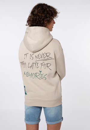 Frau trägt beige Kapuzenpullover mit hochgezogener Kapuze, Text "Es ist niemals zu spät für Erinnerungen" auf dem Rücken, kombiniert mit hochgekrempelten hellblauen Jeansshorts.