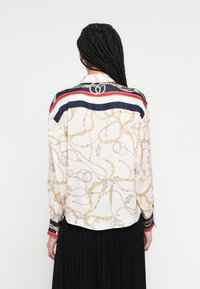 Košulja dugih rukava u krem boji s uzorkom zlatnog lanca. Tamnoplavi i crveni prugasti detalji na ovratniku i manžetama. Mekani, lagani materijal.