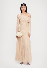 Beige één-schouder jurk met gerimpeld stof, doorschijnende overlay en pailletten. Vloeiende tule rok. Model houdt een witte, ronde handtas vast. Zwarte sandalen.