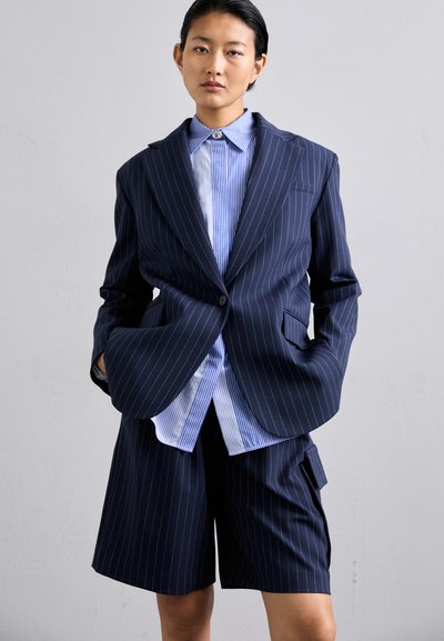 Veste de costume bleu marine à rayures, accompagnée de shorts assortis, superposée sur une chemise à rayures bleues et blanches. Tissu lisse et coupe ajustée.