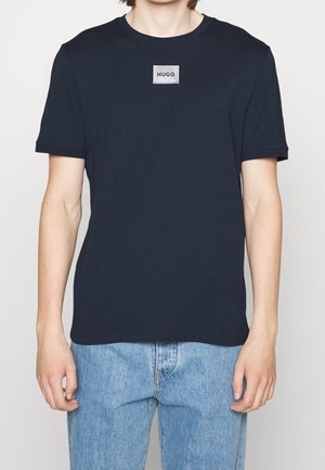 Mann trägt ein schlichtes schwarzes T-Shirt der Marke Hugo und hellblaue Jeans, steht mit entspannten Armen seitlich vor einem einfarbigen Hintergrund.