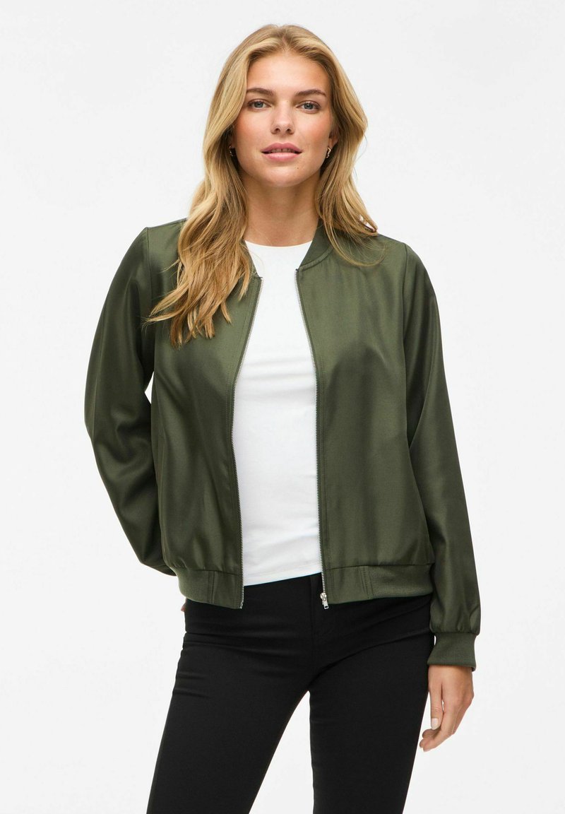 Giacca bomber verde oliva in tessuto satinato, chiusura con zip, polsini e orlo a coste, indossata sopra un top bianco e abbinata a pantaloni neri.