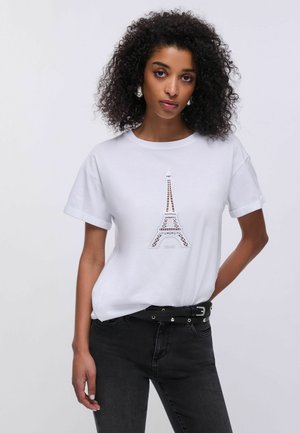 Femme aux cheveux bouclés portant un t-shirt blanc avec un motif de la tour Eiffel, un jean noir et une ceinture noire à double boucle, debout devant un fond uni.