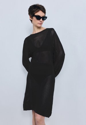 Γυναίκα που φοράει μακρυμάνικο μαύρο φόρεμα από δίχτυ και μαύρα γυαλιά ηλίου, όρθια μπροστά σε ένα απλό ανοιχτό γκρι φόντο.