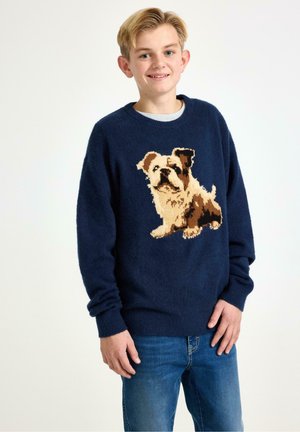 Maglione blu navy con design lavorato a maglia e grafica di un cane in crema e marrone, abbinato a jeans blu.