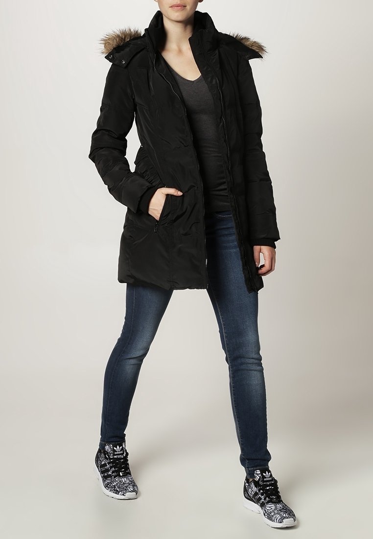 Femme portant un manteau d'hiver noir à capuche avec bordure en fourrure, un t-shirt gris foncé, un jean bleu et des baskets Adidas noir et blanc, debout avec une main dans la poche.