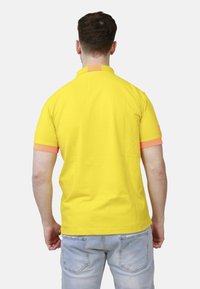 Uomo in camicia polo gialla con polsini arancioni e jeans blu sbiaditi, in piedi e girato di spalle contro uno sfondo semplice.