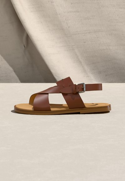 Καφέ δερμάτινο σανδάλι με επίπεδη σόλα, λουριά που διασταυρώνονται και ασημί αγκράφα, πάνω σε ελαφρώς μπεζ επιφάνεια με φόντο ύφασμα.