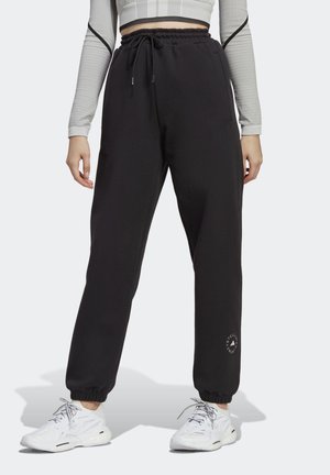 Mujer con joggers negros con cordón y puños en los tobillos y zapatillas blancas, de pie frente a un fondo liso.