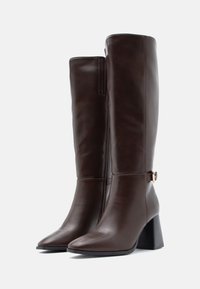 Hnědé kožené vysoké boty ke kolenům s špičatou špičkou, blokovým podpatkem a bočním zipem. Obsahují pásek a kovový akcent. Hladký povrch.