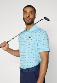 Polo shirt albastru deschis cu un model de stele negre, cu mâneci scurte și guler, asociat cu pantaloni bleumarin și un curea asortată.