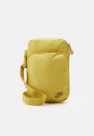 Żółta nylonowa torba na ramię o prostokątnym kształcie, z regulowanym paskiem, zapięciem na zamek błyskawiczny oraz brązowym logo Nike na przodzie.