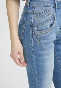 Jeans di denim blu chiaro con una texture leggermente sbiadita, caratterizzati da design unici delle tasche, bottoni in metallo e dettagli di cuciture intricate.