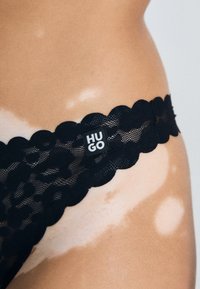 String noir en dentelle avec une taille festonnée et une petite étiquette "HUGO" sur le côté. La dentelle présente un motif floral.