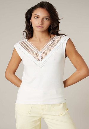 Vrouw met donker haar, gekleed in een mouwloos wit topje met een kanten V-hals en lichtbeige broek, staand met haar handen achter haar rug.