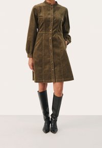 Robe en velours côtelé kaki avec manches longues bouffantes, fermeture par boutons, couture à la taille et poches latérales, associée à des bottes noires montantes jusqu'aux genoux.