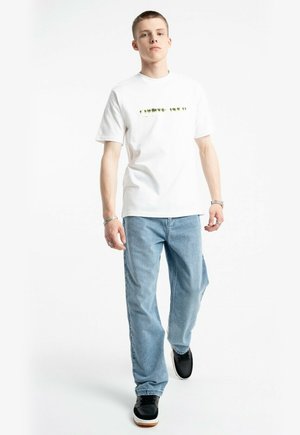 Młody mężczyzna idzie do przodu, ubrany w białą koszulkę z grafiką, jasnoniebieskie dżinsy, czarne sneakersy i srebrne bransoletki na obu nadgarstkach.